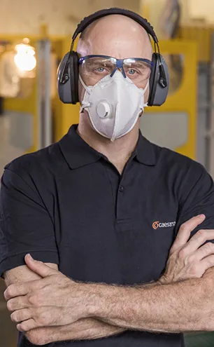 Fabricator wearing protective gear including a respirator mask, safety glasses, and earmuffs, standing confidently in a Caesarstone workshop.