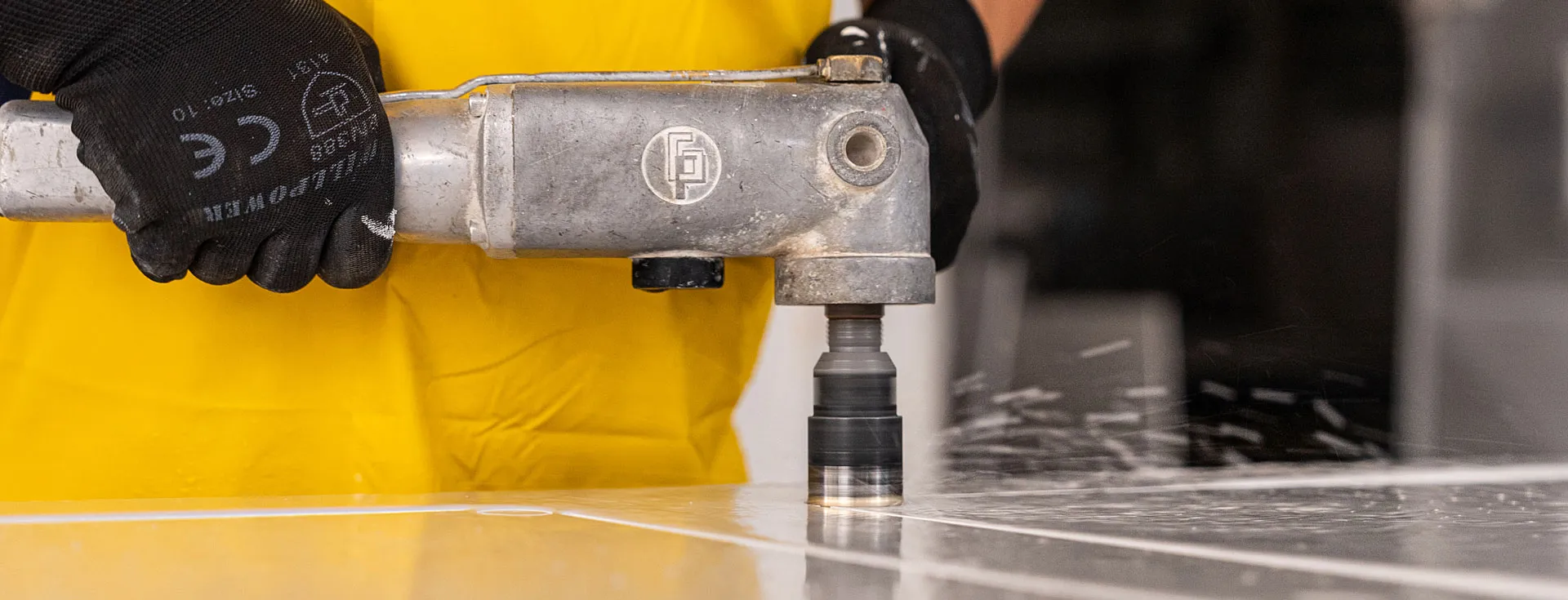 About Caesarstone Master of Stone - a fabricator wearing gloves and yellow apron using a handheld drill on a flat surface.